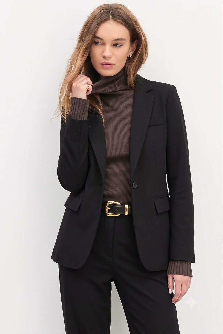 Woman wearing The Worth Blazer Black styled over a brown ribbed turtleneck with black tailored pants and a gold-buckled belt, showcasing a timeless minimalist and versatile workwear look.