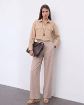 A model wearing The Portofino Linen Set featuring a minimalist cropped beige linen jacket with front pockets and high-waisted wide-leg linen trousers, styled with neutral sandals and a textured dark brown clutch, embodying refined basics and versatile staples for a timeless capsule wardrobe.