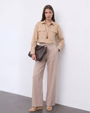 A model wearing The Portofino Linen Set featuring a minimalist cropped beige linen jacket with front pockets and high-waisted wide-leg linen trousers, styled with neutral sandals and a textured dark brown clutch, embodying refined basics and versatile staples for a timeless capsule wardrobe.