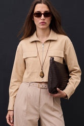 Woman wearing The Portofino Linen Set in neutral tones with a cropped jacket and high-waisted trousers, paired with dark sunglasses and holding a textured brown clutch against a black background.