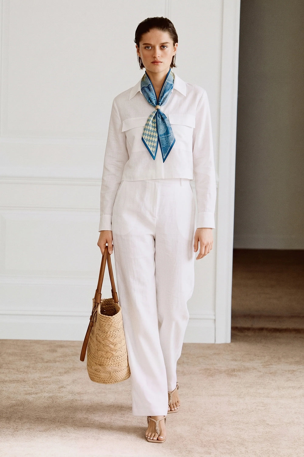 Woman wearing The Santorini Linen Shirt – Ivory with white pants, styled with a blue patterned scarf, carrying a woven tote bag in a minimalist, timeless room setting.