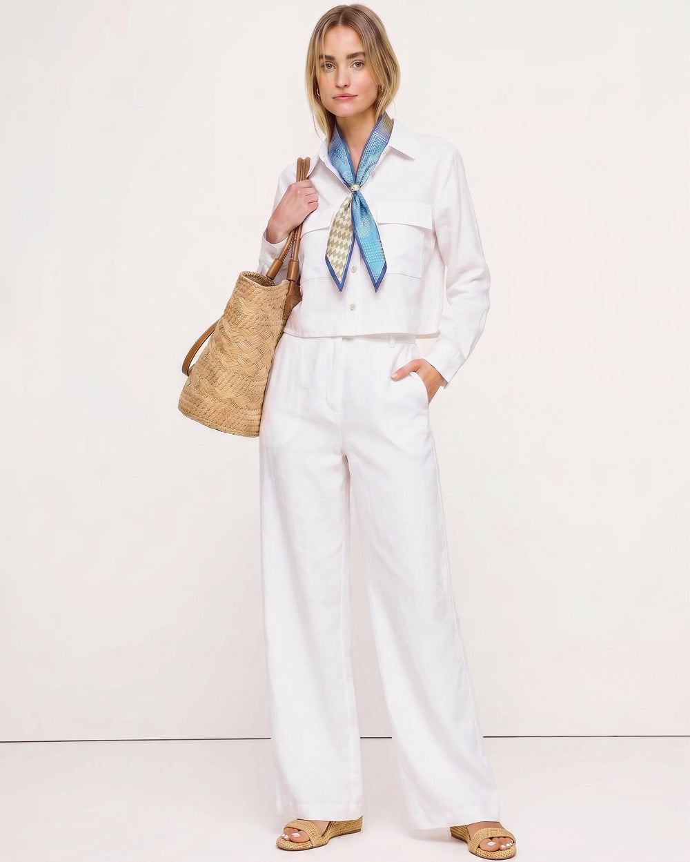 Woman wearing The Santorini Linen Shirt – Ivory styled with wide-leg linen pants, a blue patterned scarf, woven tote bag, and sandals showcasing a timeless, minimalist capsule wardrobe look.