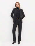 A confident woman wearing The Worth Blazer Black styled with tailored black pants, a black turtleneck, layered gold necklaces, a black belt with a gold buckle, and black pointed-toe boots, embodying timeless minimalist workwear.