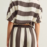 Back view of a model wearing The Miou Muse Boat Neck Stripe Top with wide horizontal brown and cream stripes, featuring a boat neckline and relaxed fit, styled with matching striped pants.
