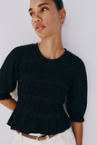 Woman wearing The Vivienne Peplum Blouse in black, featuring delicate ruched detailing and puffed sleeves, styled with white trousers for a modern capsule wardrobe look.