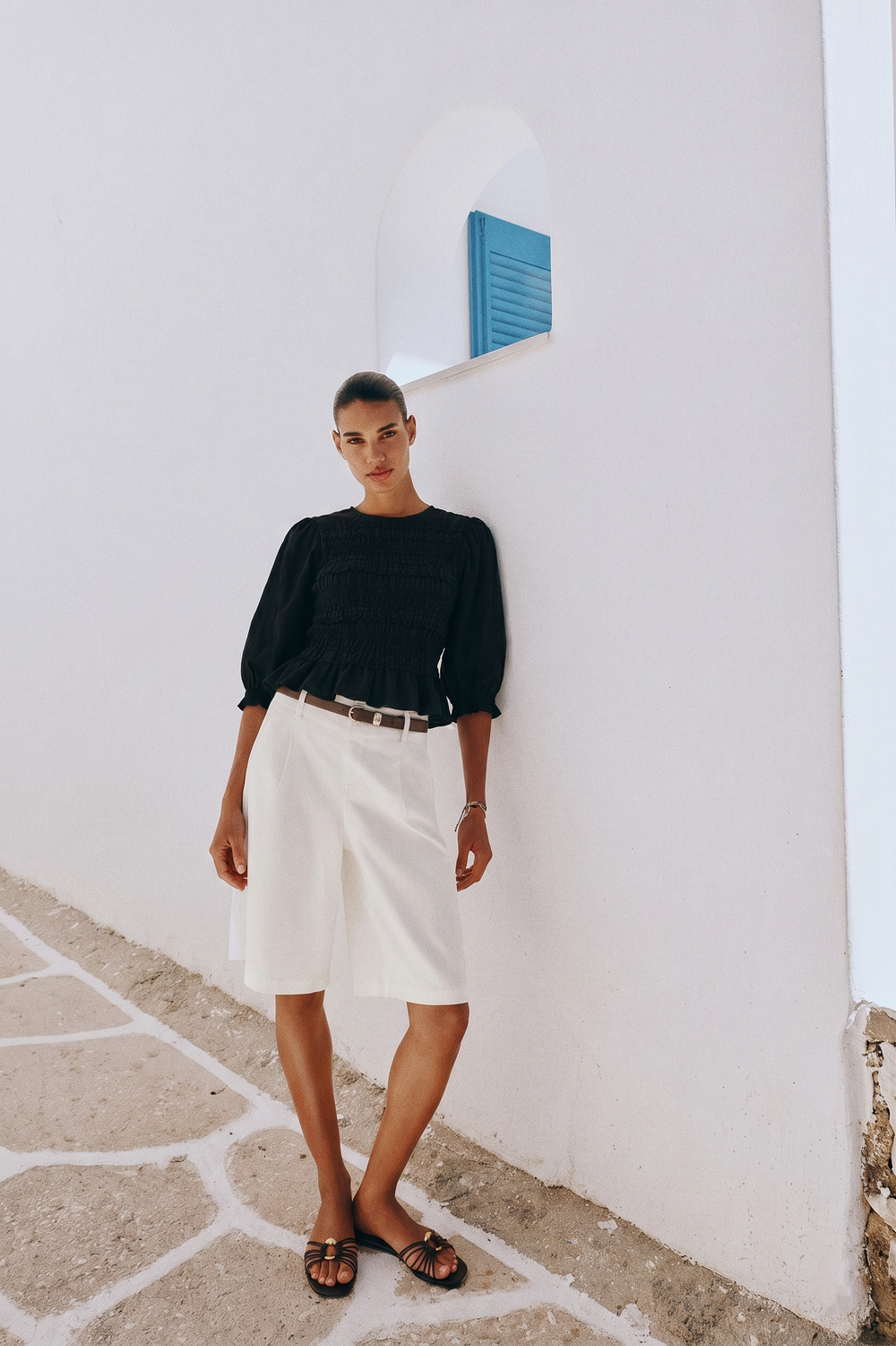 Woman wearing The Vivienne Peplum Blouse in black, styled with white tailored shorts and black sandals, standing against a white wall with minimalist architecture.