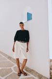 Woman wearing The Vivienne Peplum Blouse in black, styled with white tailored shorts and black sandals, standing against a white wall with minimalist architecture.