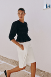 Model wearing The Vivienne Peplum Blouse in black, featuring a smocked bodice and peplum hem, styled with white tailored shorts for a refined, modern capsule wardrobe look.
