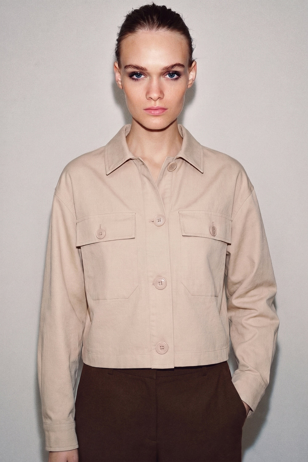 Woman wearing The Sahara Linen Set featuring a minimalist, clean-lined beige linen jacket with button closures and chest pockets, paired with dark brown trousers, styled with a neutral background.