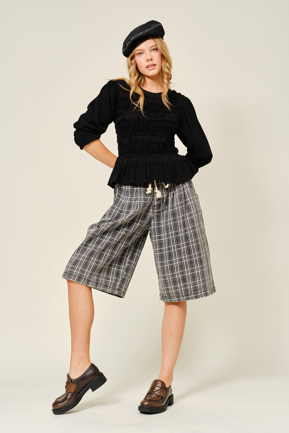 Model wearing The Vivienne Peplum Blouse in black with smocked detailing and peplum hem, paired with plaid wide-leg shorts, leather loafers, and a black beret for a modern capsule wardrobe look.