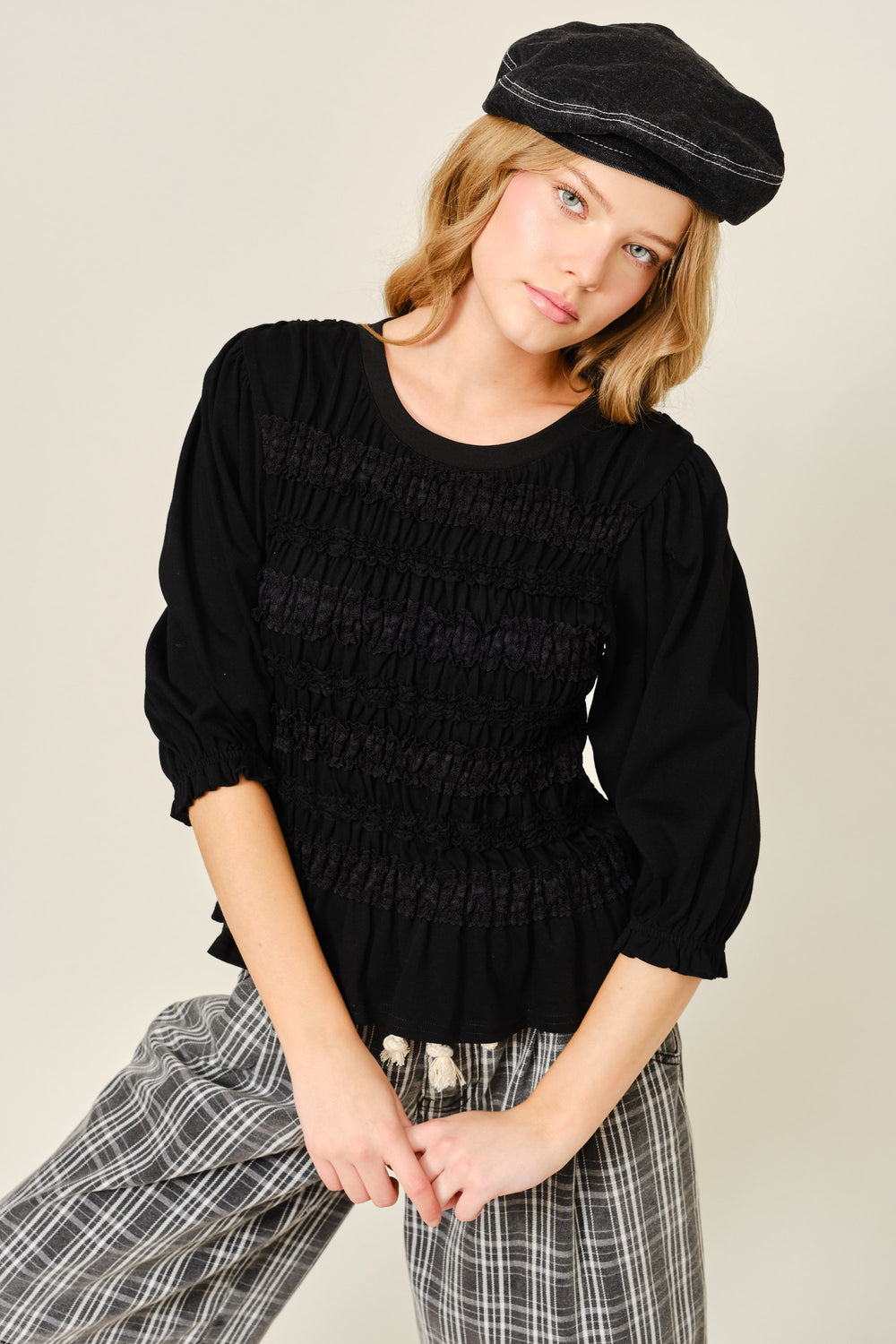 Young woman wearing The Vivienne Peplum Blouse in black with ruched detailing, paired with relaxed plaid pants and a casual black cap, styled for a modern minimalist look.