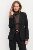 Woman wearing The Worth Blazer Black styled over a brown ribbed turtleneck with black tailored pants and a gold-buckled belt, showcasing a timeless minimalist and versatile workwear look.