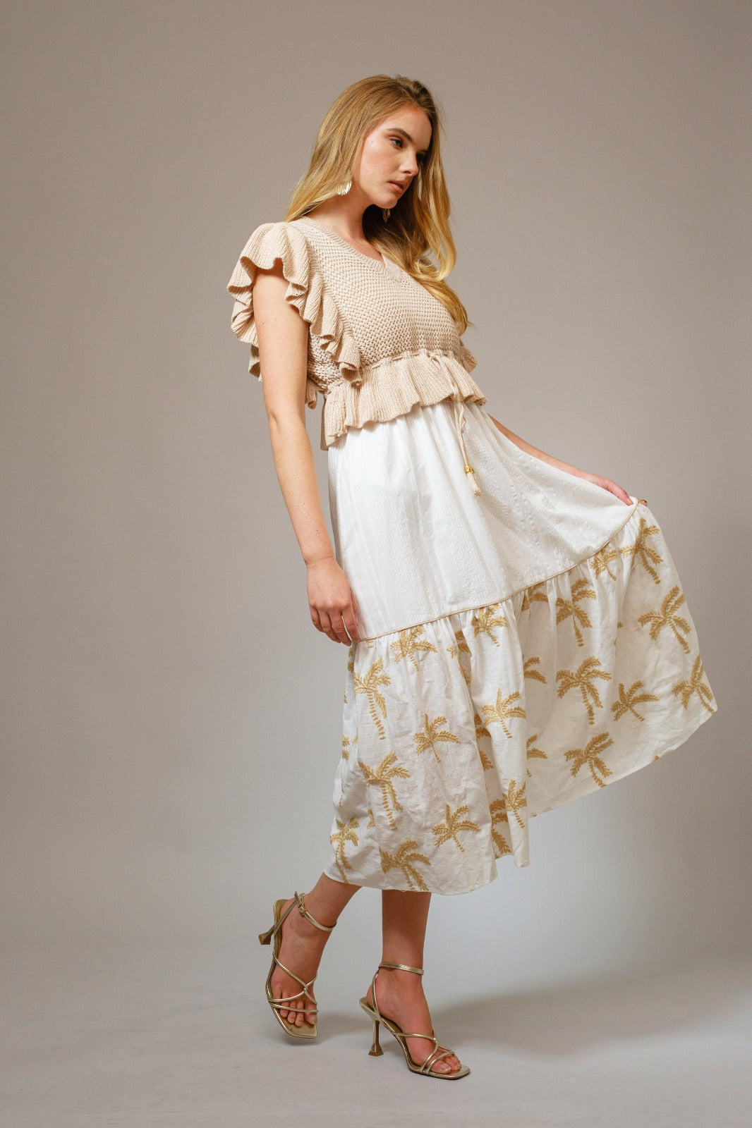 Woman wearing The Coco Palm Dress featuring a white flowing skirt with subtle palm tree patterns, styled with a beige textured ruffle top and strappy heels, embodying timeless minimalism.