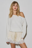 A woman wearing "The Camille Lace Satin Short" in a soft cream satin with delicate lace trim, paired with a loose off-shoulder white sweater, showcasing a modern, feminine, and versatile capsule wardrobe piece.