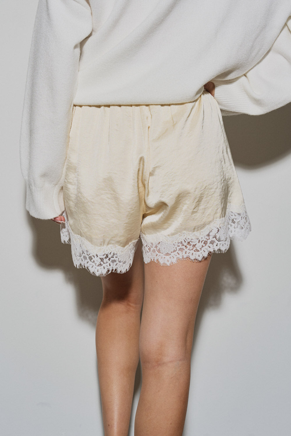 Rear view of a model wearing The Camille Lace Satin Short in a soft cream color, featuring stretchable fabric and delicate lace trim along the hem, styled with a white sweater.