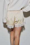 Rear view of a model wearing The Camille Lace Satin Short in a soft cream color, featuring stretchable fabric and delicate lace trim along the hem, styled with a white sweater.