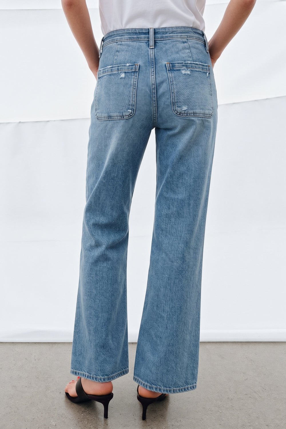Back view of a person wearing light blue denim Camden Ankle Jean with a relaxed fit, slightly distressed patch pockets, paired with black heeled sandals and a white top.