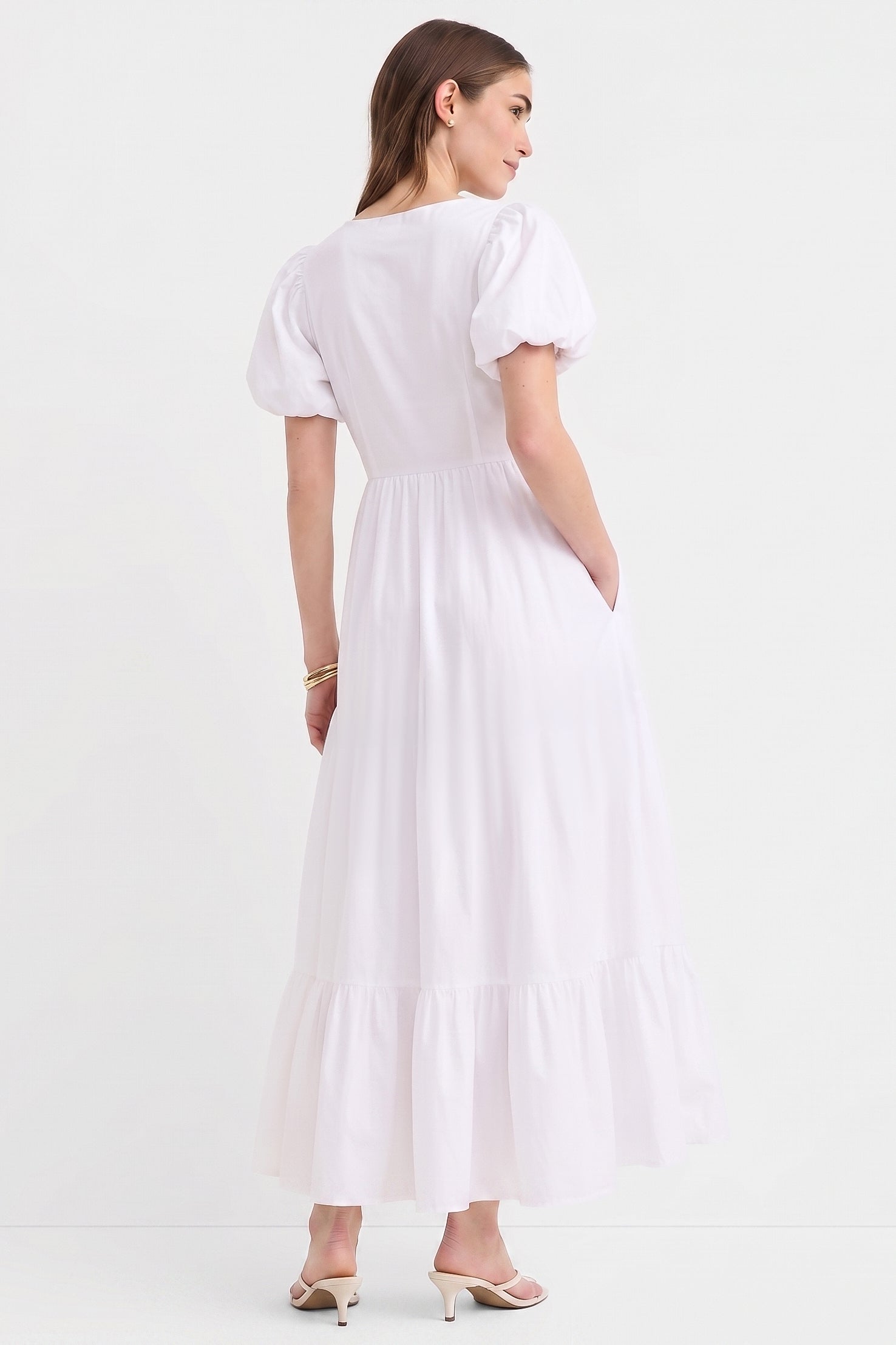 Woman wearing The Belle Maxi Dress in white, featuring puff sleeves, a gathered waist, stretchable fabric, and a flowing tiered hem, styled with minimalist heels.