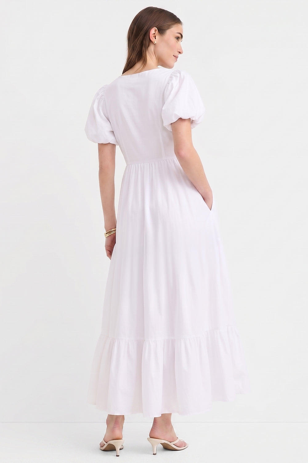 Woman wearing The Belle Maxi Dress in white, featuring puff sleeves, a gathered waist, stretchable fabric, and a flowing tiered hem, styled with minimalist heels.