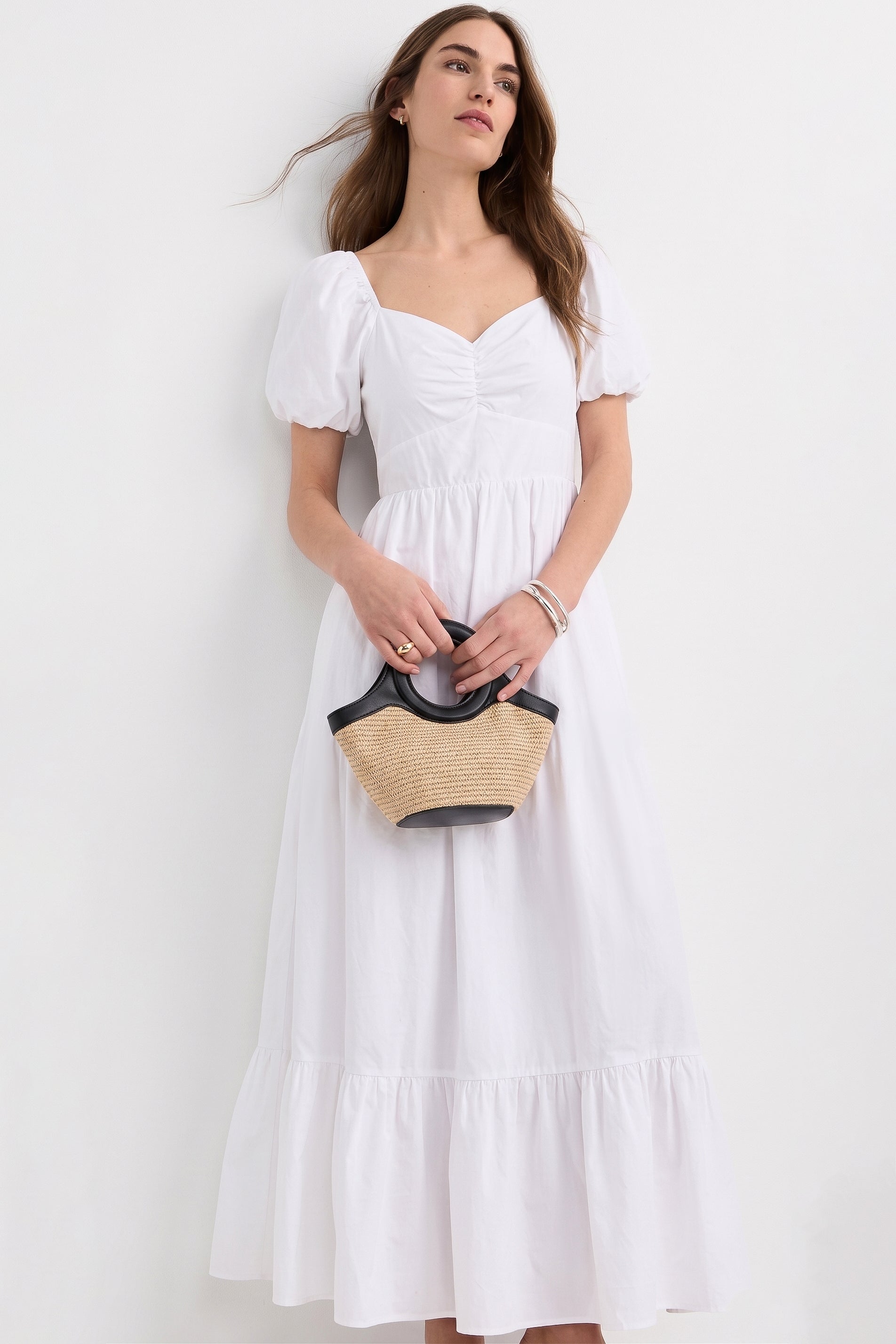 Woman wearing The Belle Maxi Dress in white with puff sleeves and a gathered bodice, holding a small straw handbag with black handles, styled for a timeless capsule wardrobe look.