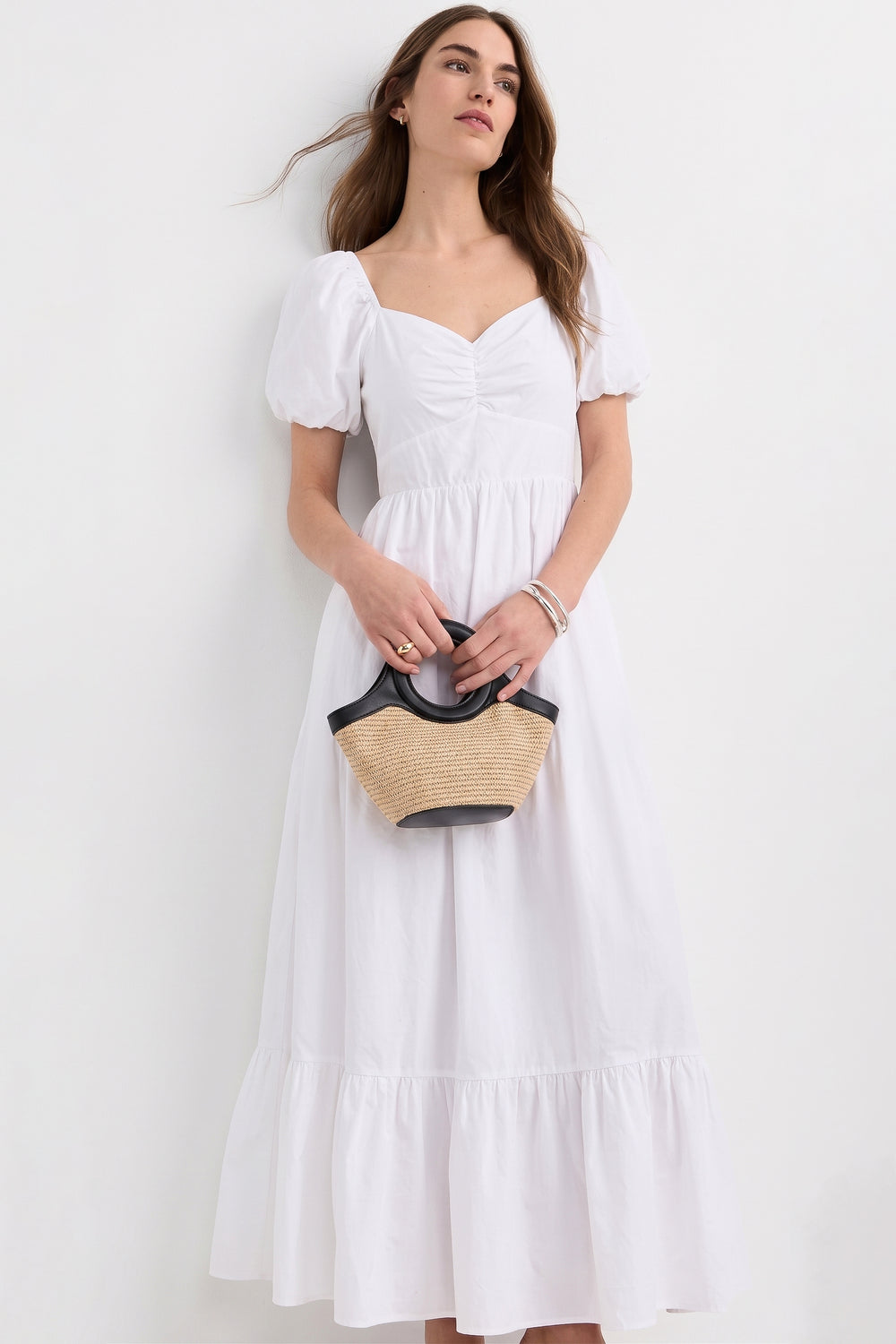 Woman wearing The Belle Maxi Dress in white with puff sleeves and a gathered bodice, holding a small straw handbag with black handles, styled for a timeless capsule wardrobe look.