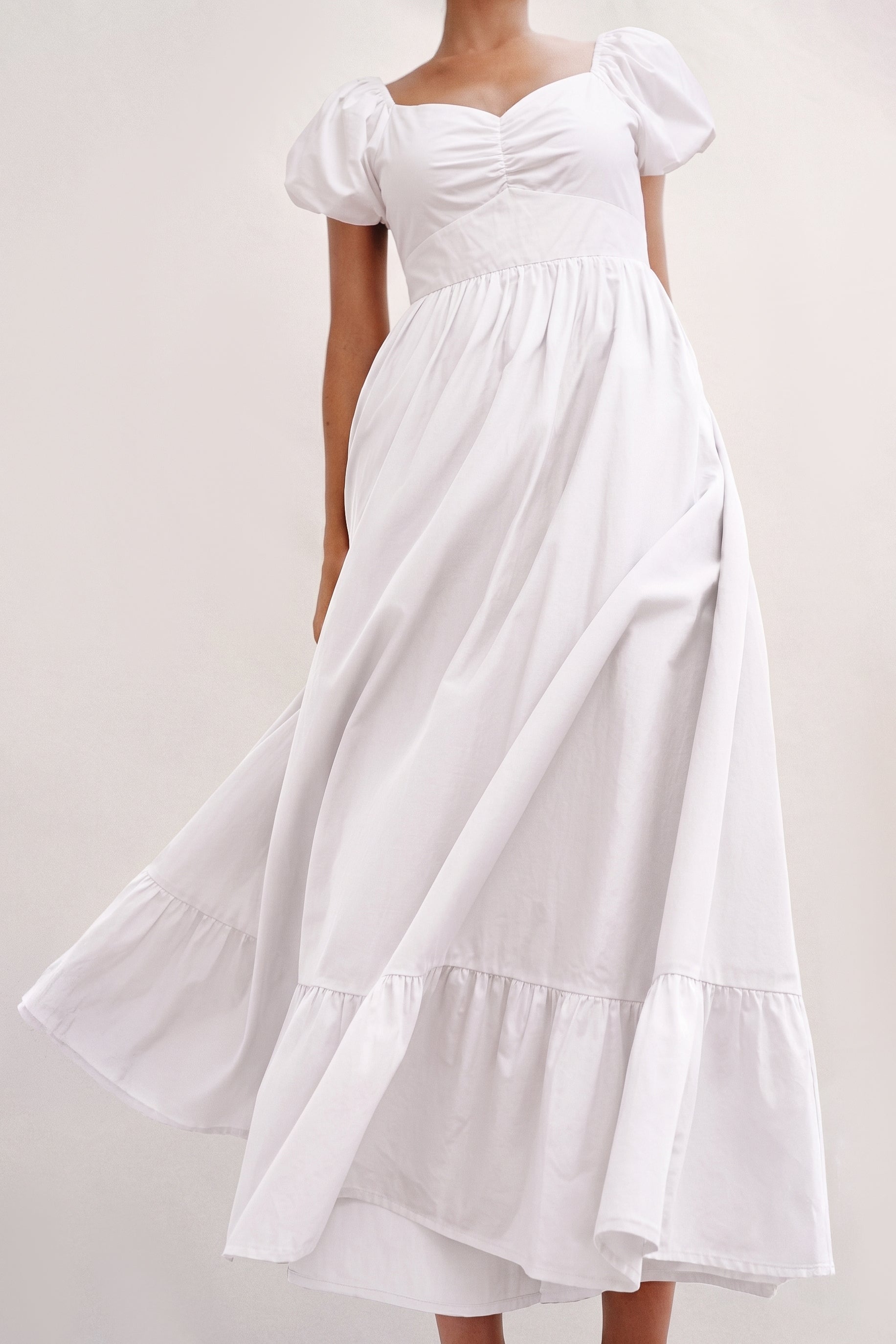 A woman wearing The Belle Maxi Dress in white, featuring stretchable fabric, puff sleeves, and a gathered bodice with a flowing, tiered skirt, embodying timeless minimalism.
