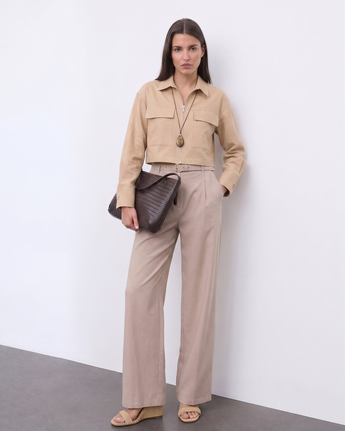 A model wearing The Portofino Linen Set featuring a minimalist cropped beige linen jacket with front pockets and high-waisted wide-leg linen trousers, styled with neutral sandals and a textured dark brown clutch, embodying refined basics and versatile staples for a timeless capsule wardrobe.
