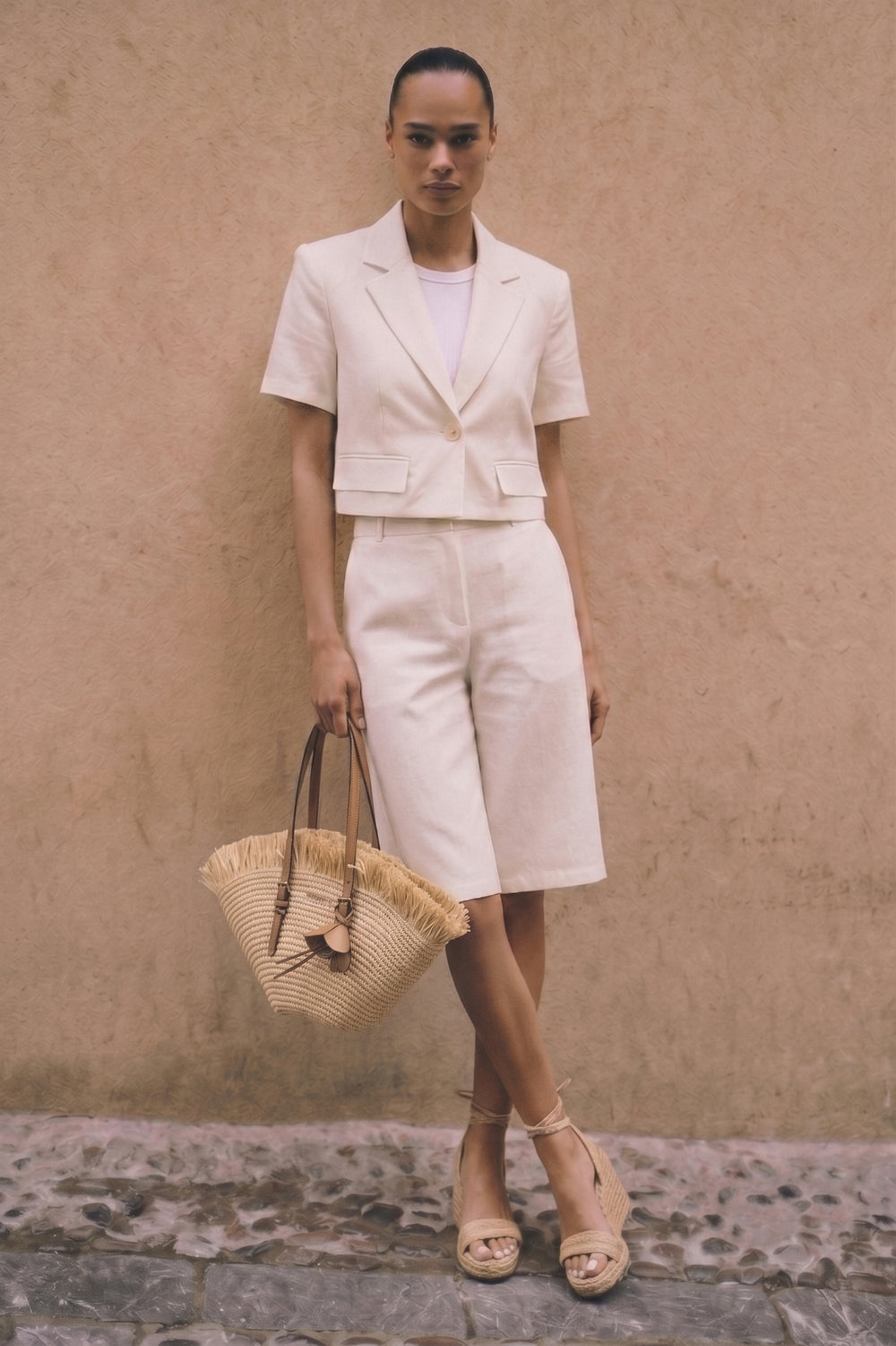 Model wearing Riviera Linen Bermuda Set featuring a cropped blazer with short sleeves and tailored Bermuda shorts, styled with neutral-toned espadrille wedges and a woven straw handbag, against a textured beige wall.
