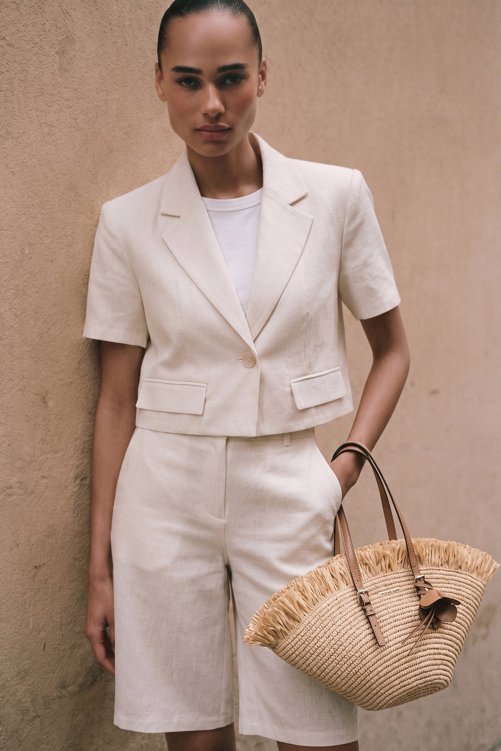 Woman wearing a tailored Riviera Linen Bermuda Set in soft neutral tones, styled with a white top and carrying a woven straw handbag, embodying timeless minimalism and refined basics.