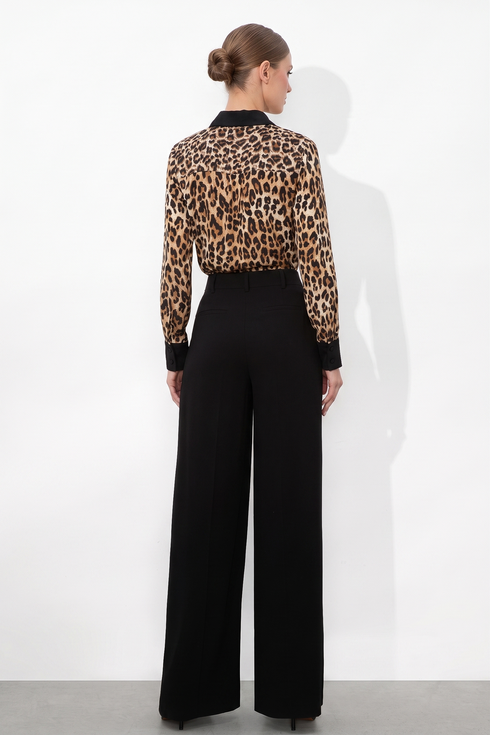 Woman standing against a white wall wearing a Leopard Print Satin Blouse with long sleeves and black cuffs, paired with high-waisted wide-leg black trousers, styled with a sleek bun hairstyle.