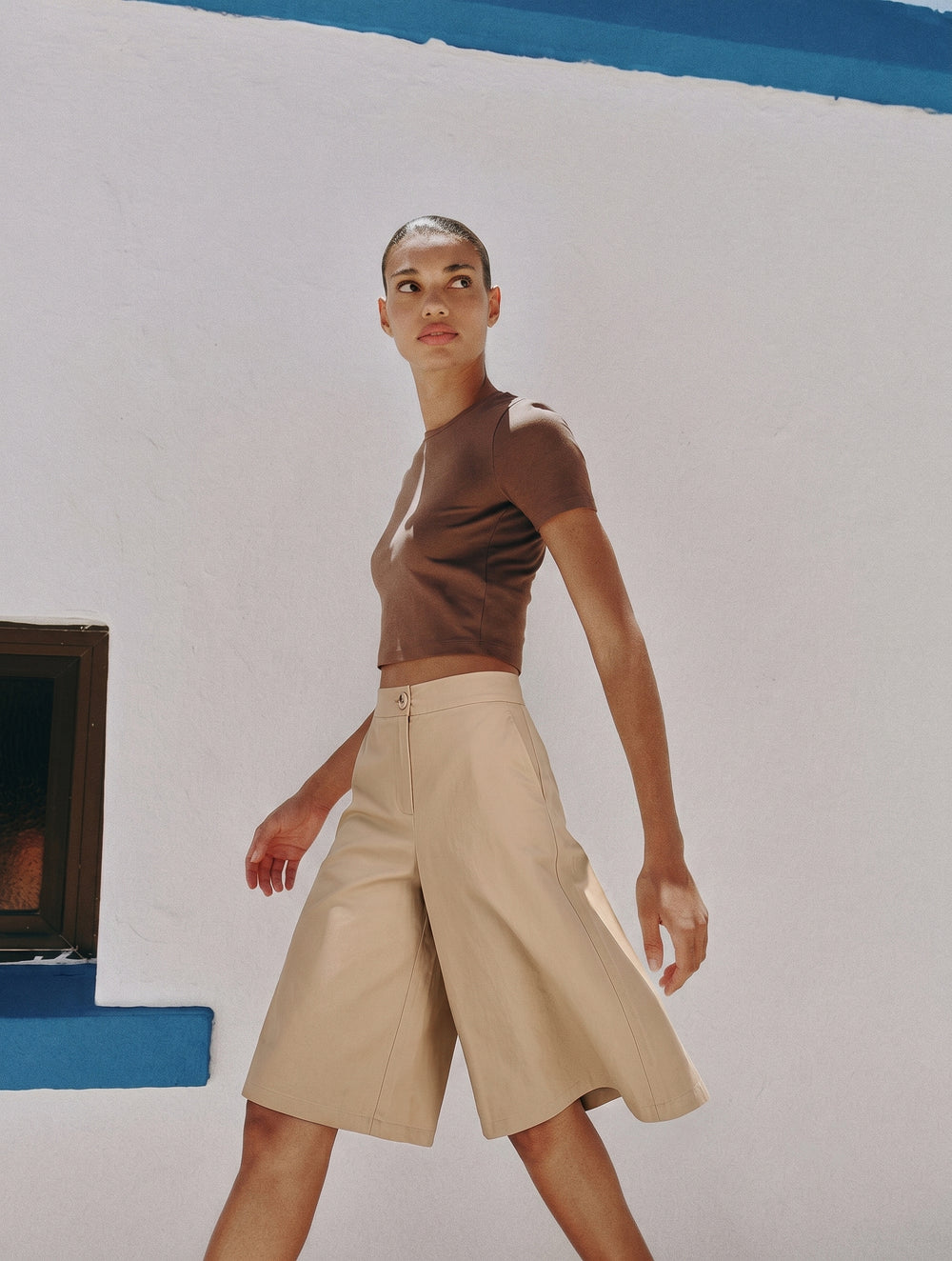 A confident woman wearing The Sahara Linen Set in neutral tones, showcasing wide-leg linen Bermuda shorts paired with a fitted minimalist brown top against a clean white and blue wall.