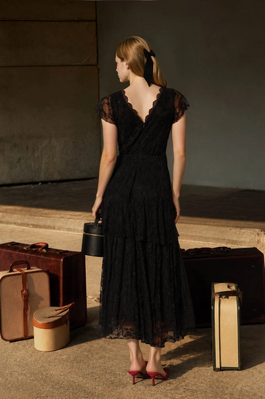 Orient Express Black Lace Maxi Dress – Whimsigoth Holiday & NYE Gown, back view of a woman in a flowing black lace gown with vintage luggage and red heels