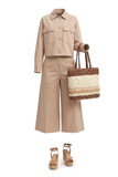 A beige two-piece outfit consisting of a cropped button-front shirt jacket and matching wide-leg pants, styled with a woven straw tote bag and tan platform sandals on a white background.