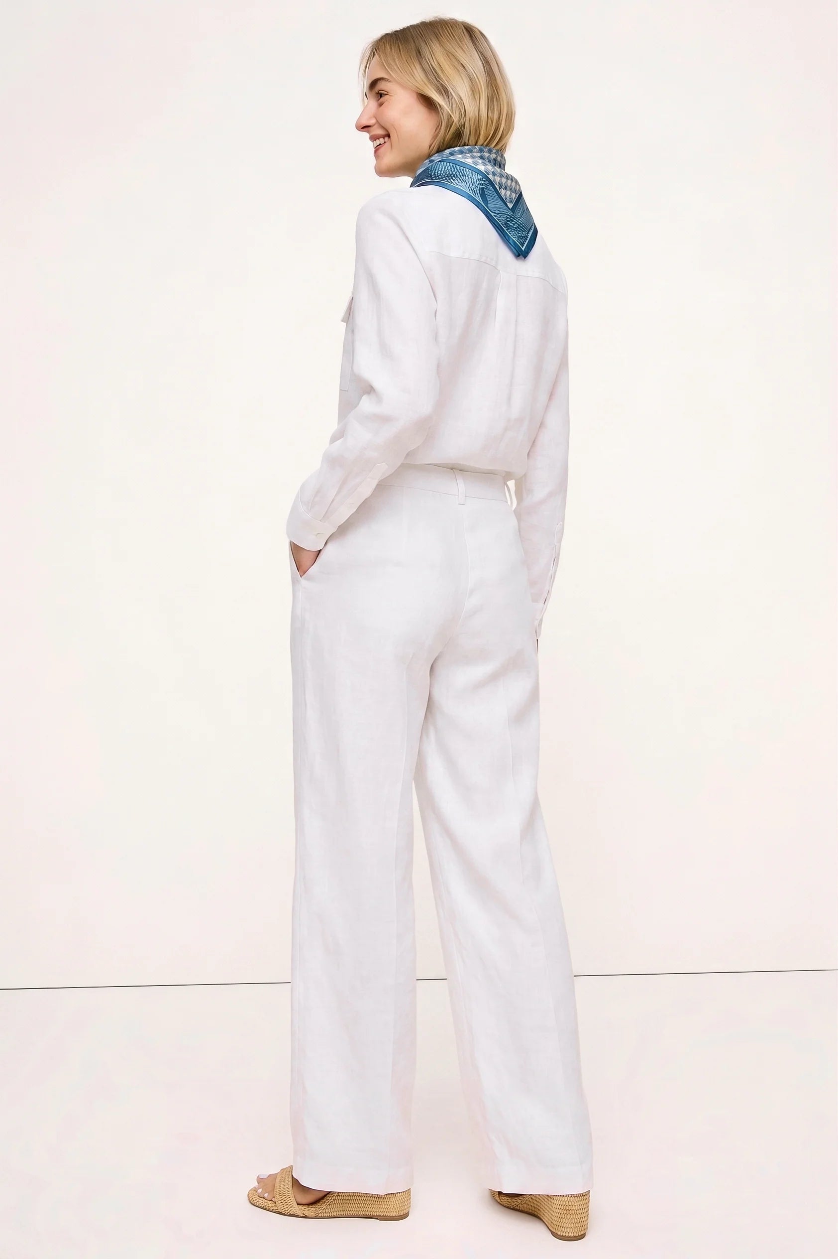 Back view of a woman wearing The Santorini Linen Shirt – Ivory paired with matching linen pants and a blue patterned scarf, showcasing a clean, timeless capsule wardrobe look.