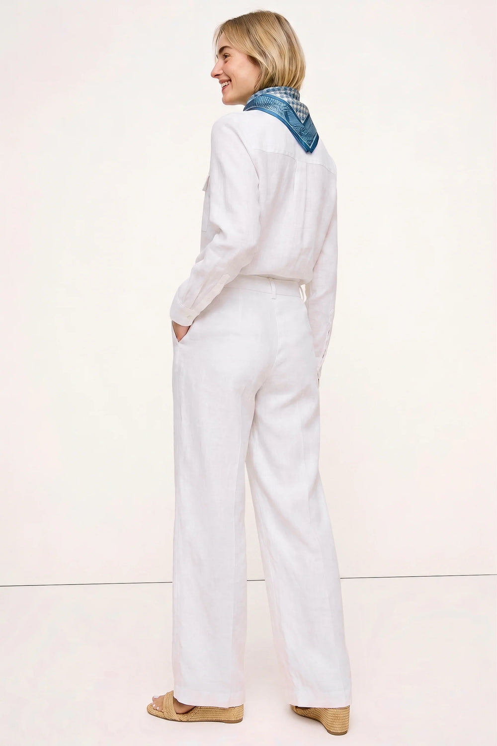Back view of a woman wearing The Santorini Linen Shirt – Ivory paired with matching linen pants and a blue patterned scarf, showcasing a clean, timeless capsule wardrobe look.