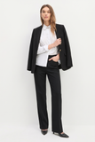 Woman wearing The Worth Blazer Black draped over her shoulders paired with tailored black pants, a white blouse with a decorative collar, and black pointed-toe heels, embodying timeless minimalist workwear.