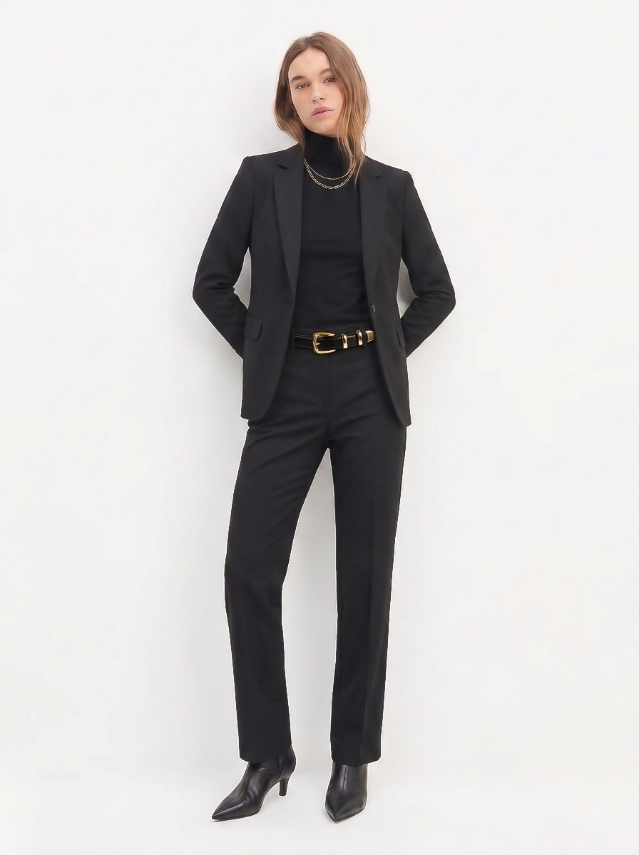 A confident woman wearing The Worth Blazer Black styled with tailored black pants, a black turtleneck, layered gold necklaces, a black belt with a gold buckle, and black pointed-toe boots, embodying timeless minimalist workwear.